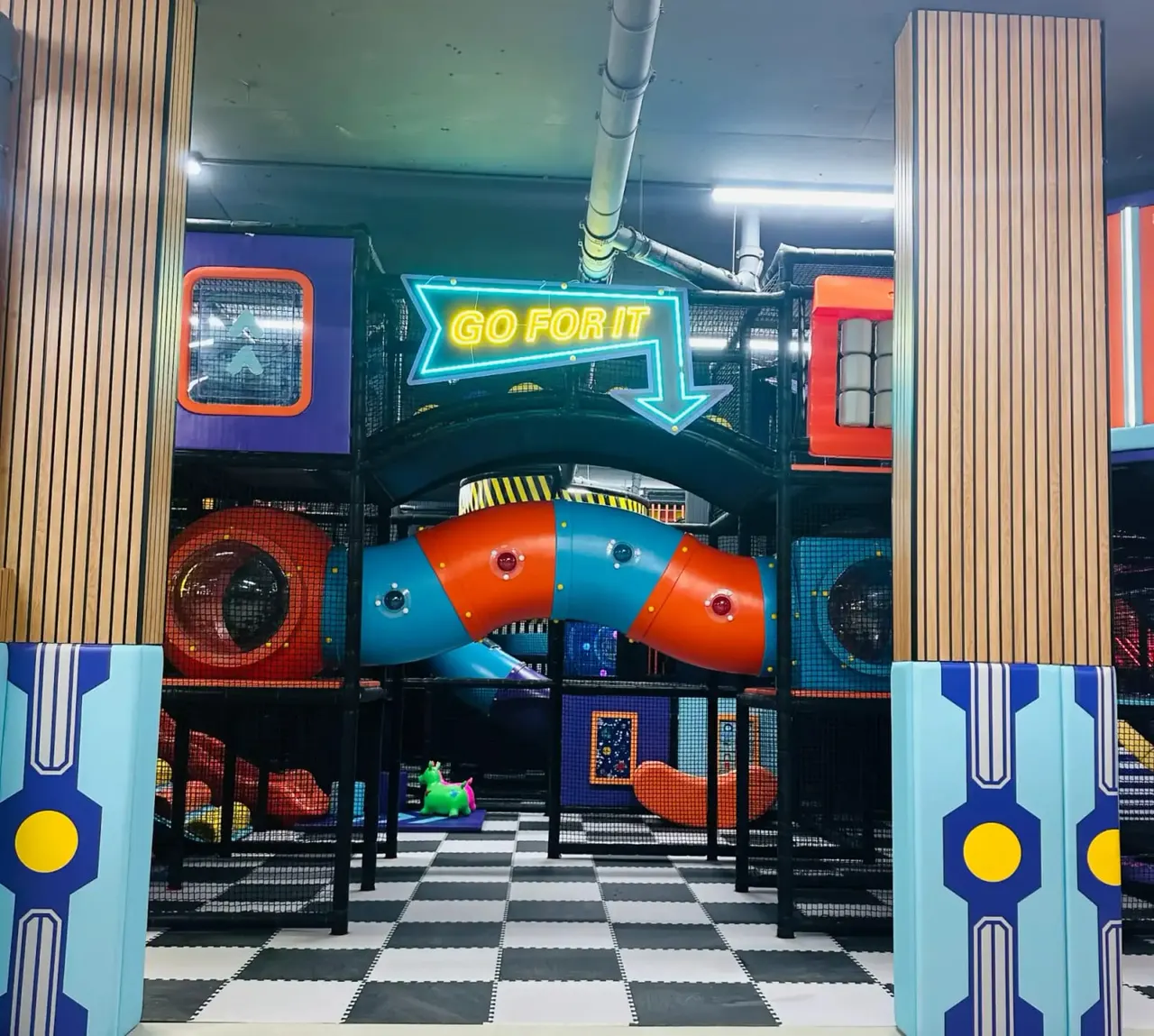 Colourful indoor soft play area with large inflatable structures in blue and orange, neon signage, and chequerboard flooring.
