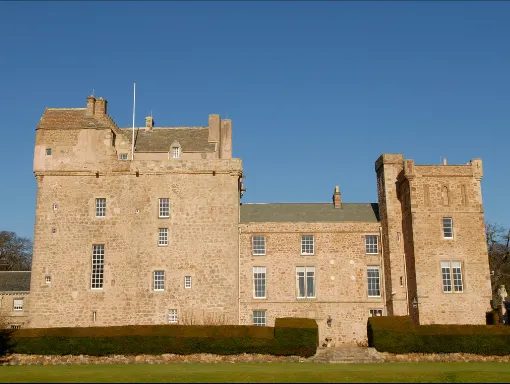 Historic stone castle with towers and crenellations set against clear blue sky, viewed across manicured lawn.