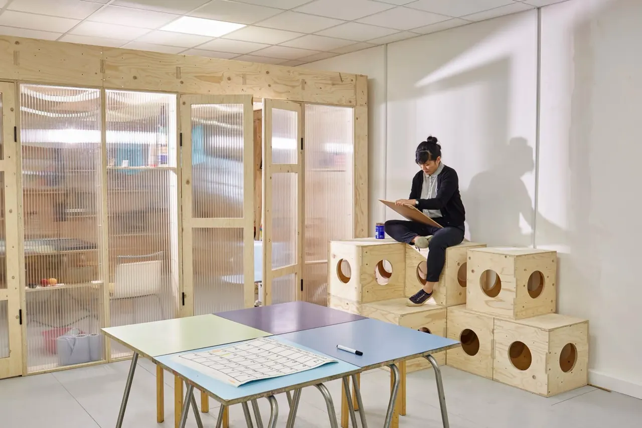 Woman sitting on stacked wooden boxes reading a clipboard in a modern activity space with modular furniture.