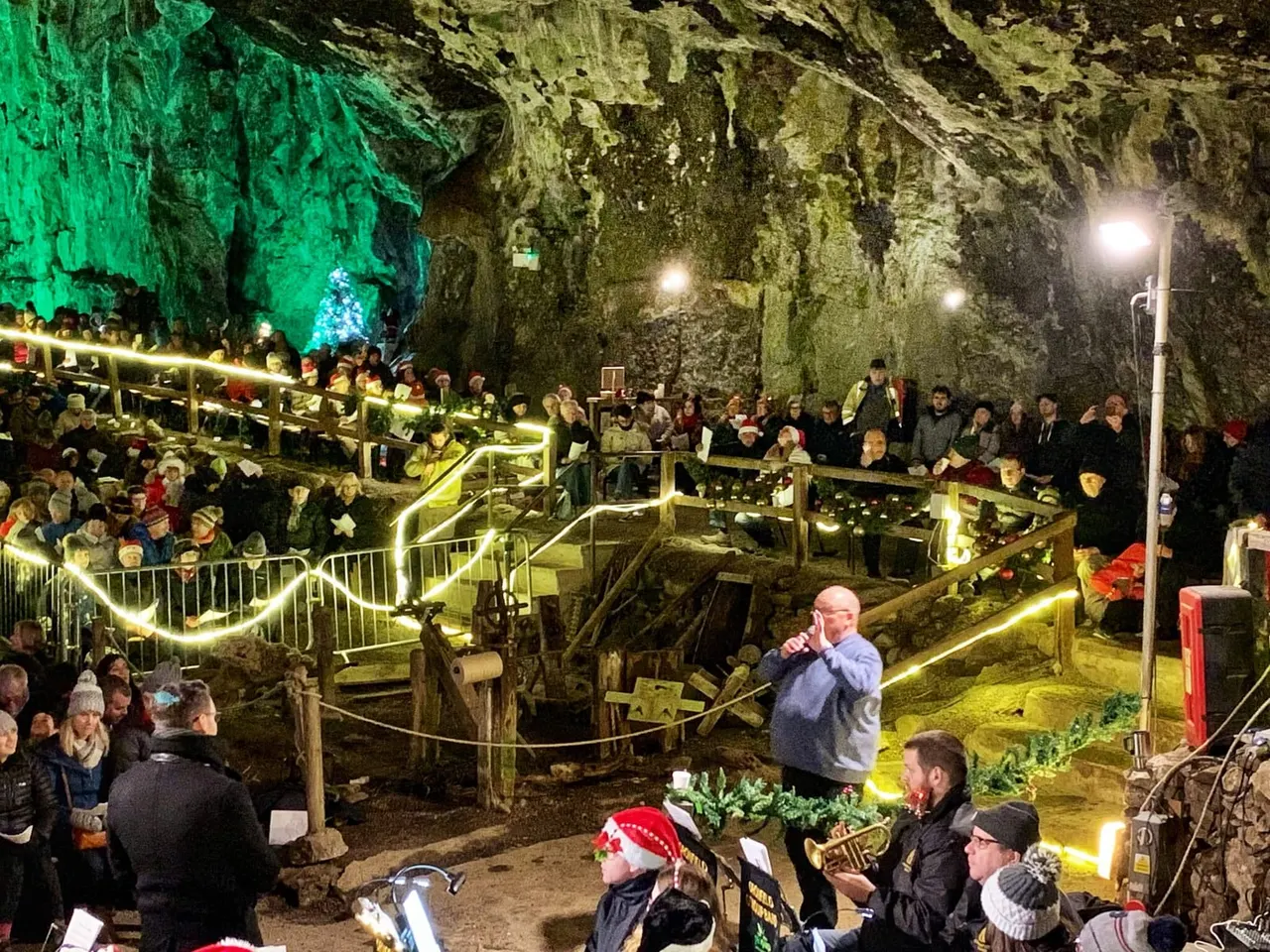 Crowds gathered inside an illuminated cave for a Christmas event, with decorated railings and a performer on stage.