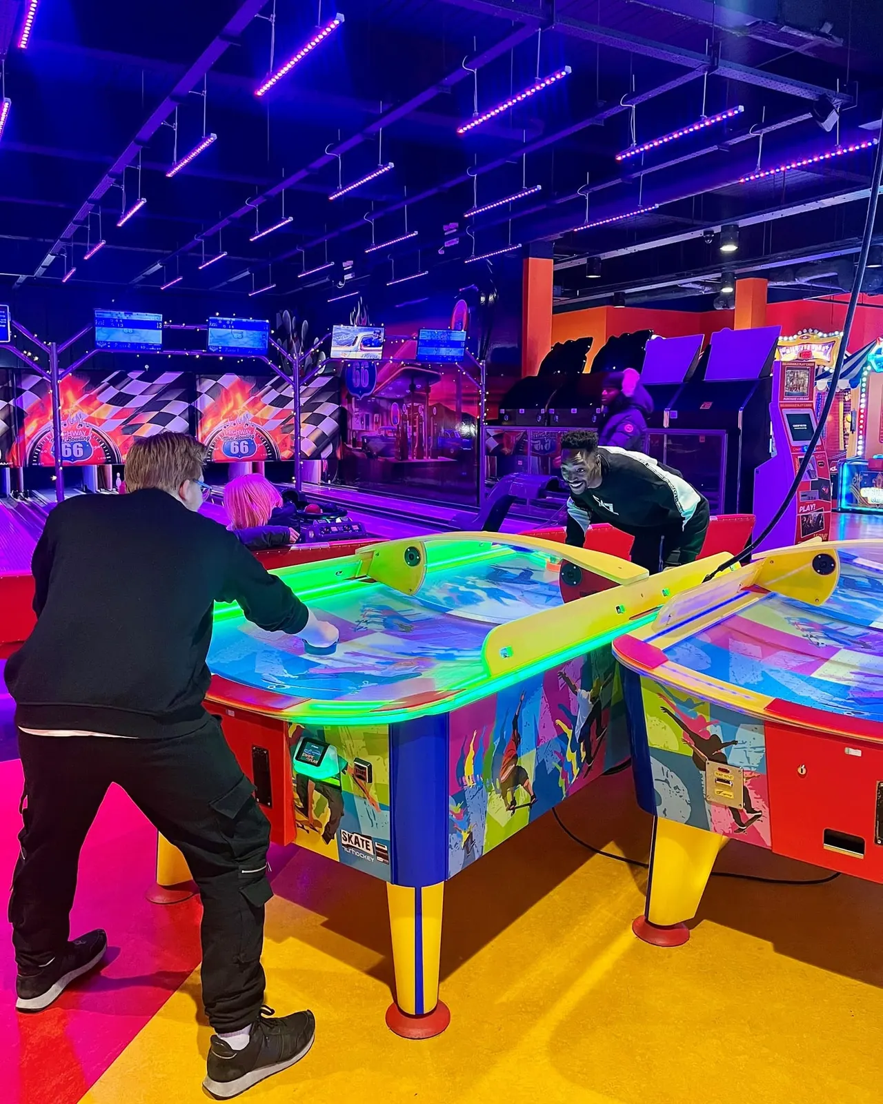 Two people playing air hockey on a neon-lit table in a vibrant arcade venue with gaming machines and colourful lighting.