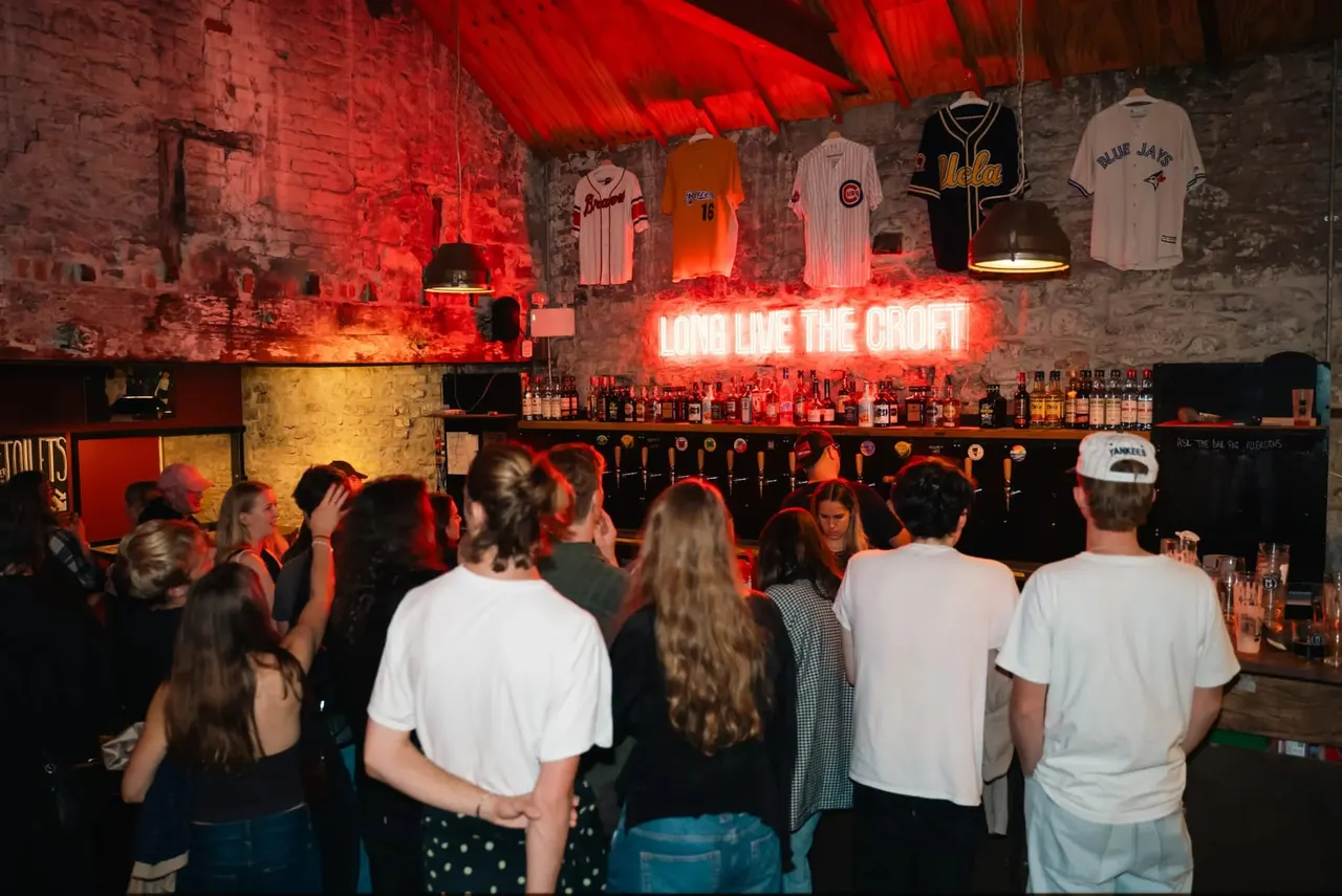 Patrons gathering at a bar beneath a neon sign, with sports jerseys displayed on the brick wall above.