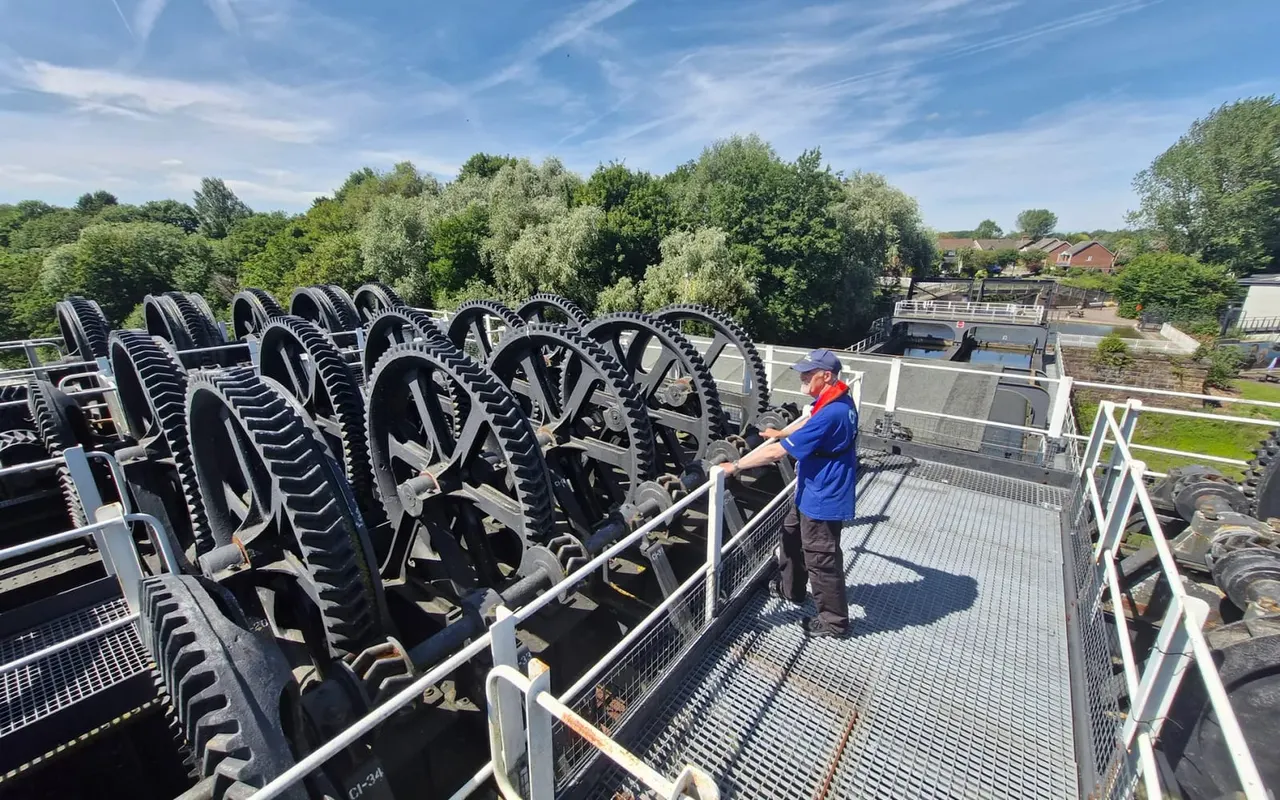A visitor in blue and red clothing stands on a viewing platform beside a row of large industrial gears at a heritage waterway site, with trees and countryside visible beyond.