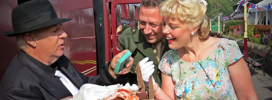Three people in vintage clothing stand beside a red heritage railway vehicle, exchanging food or refreshments.
