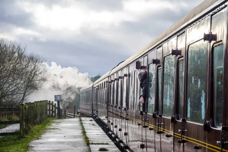 A heritage steam train with passenger carriages at a rural railway station platform, with steam rising from the locomotive in the distance.