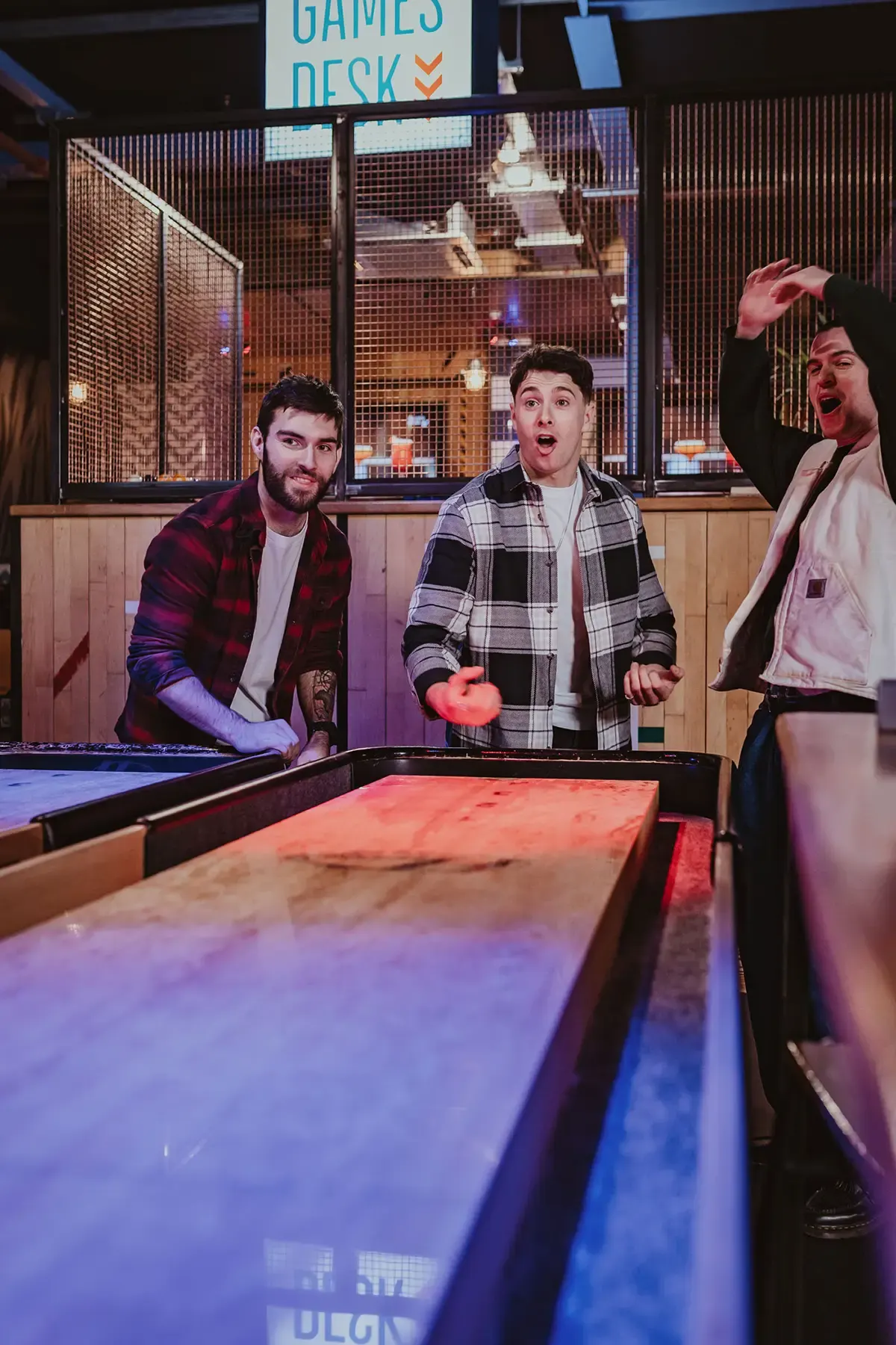 Three men playing air hockey indoors, one holding a paddle mid-game with excited expressions.