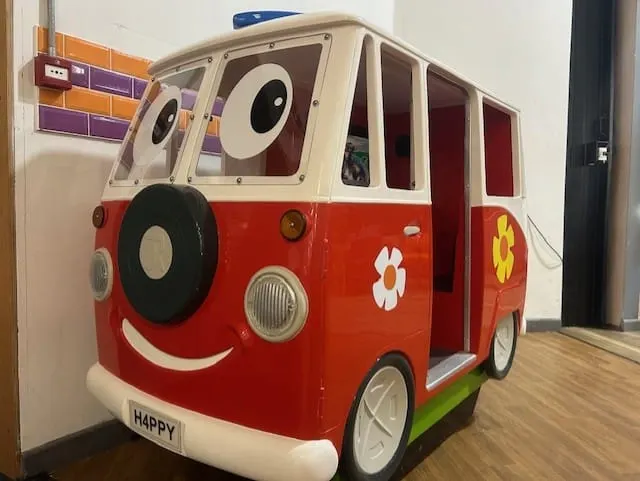 A large red and white toy bus with a friendly face, displayed indoors on wooden flooring.