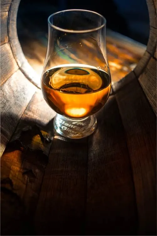 Glass of whisky on wooden barrel top, backlit by warm sunlight.
