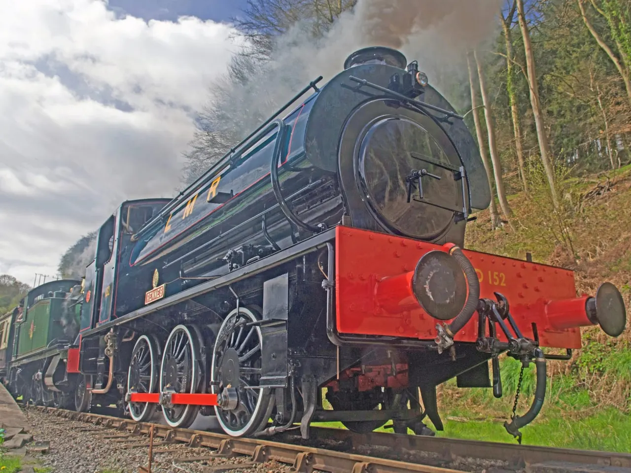 Steam locomotive with dark blue livery and red buffer beam pulling along a heritage railway line surrounded by woodland.