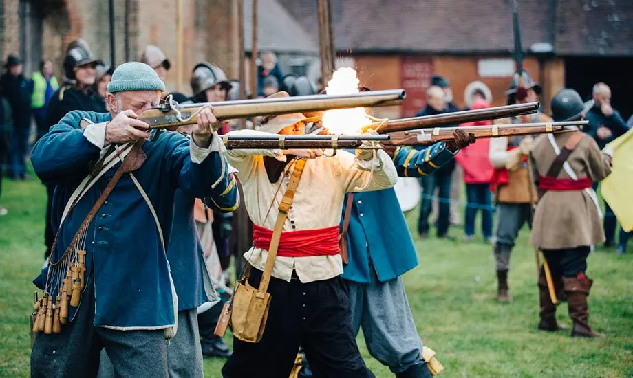 Historical reenactors firing muskets during a Civil War period demonstration, with spectators watching in the background.
