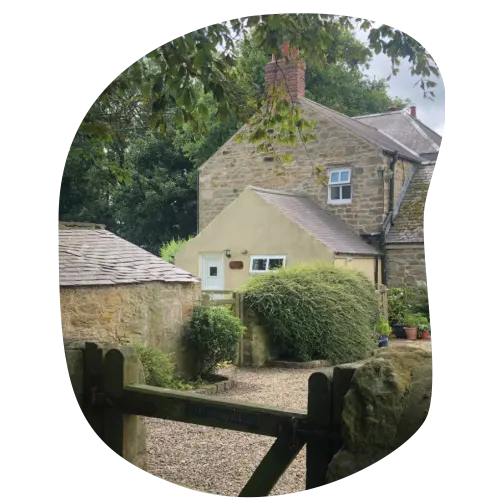 Stone cottage with cream-coloured extension surrounded by mature trees and clipped hedges, viewed through wooden gate.