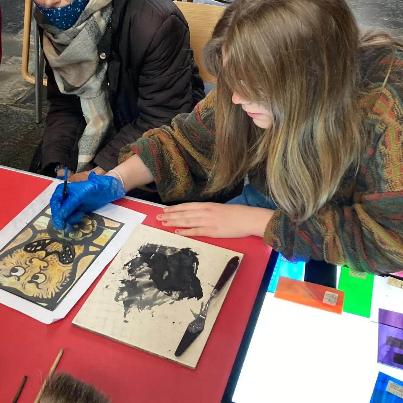 Child wearing blue gloves participating in a printmaking or art activity at a table with an adult nearby.