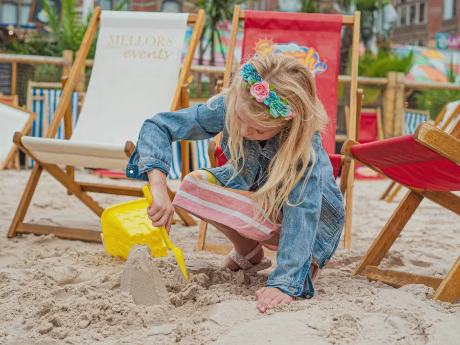 Child digging in sand with a yellow spade, surrounded by colourful deckchairs at a beach venue.
