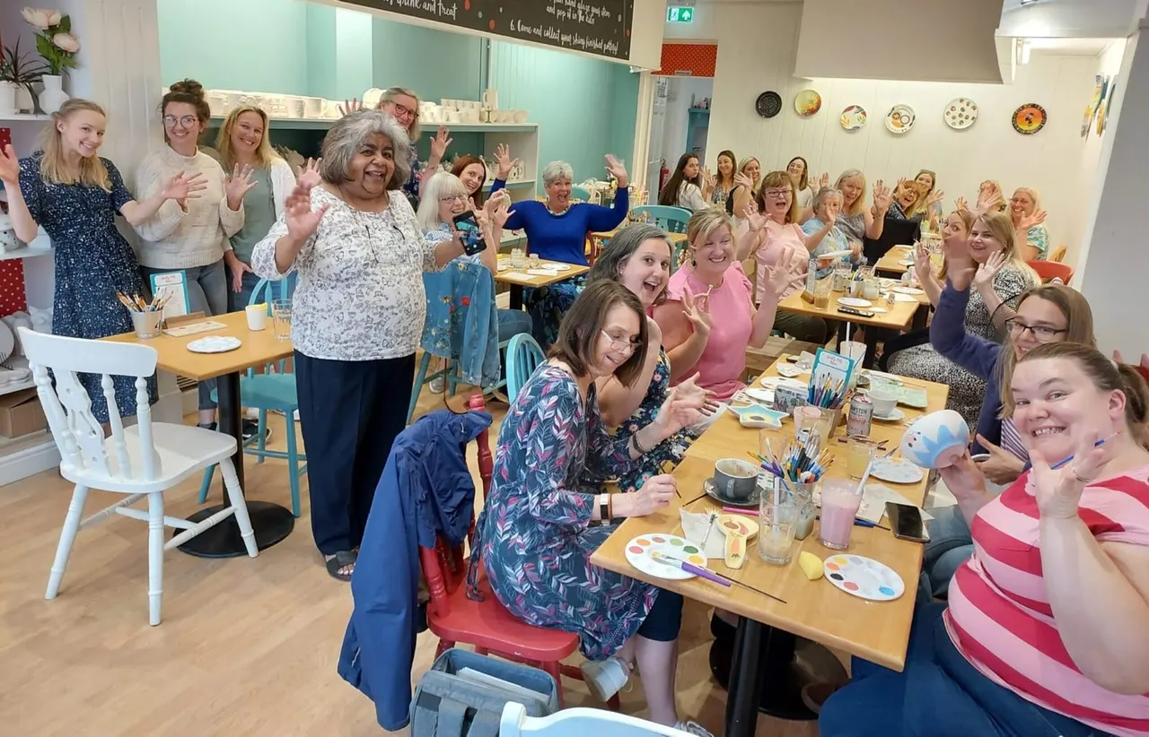 A large group of people of mixed ages waving at the camera during a pottery painting session at tables set with paints and brushes.