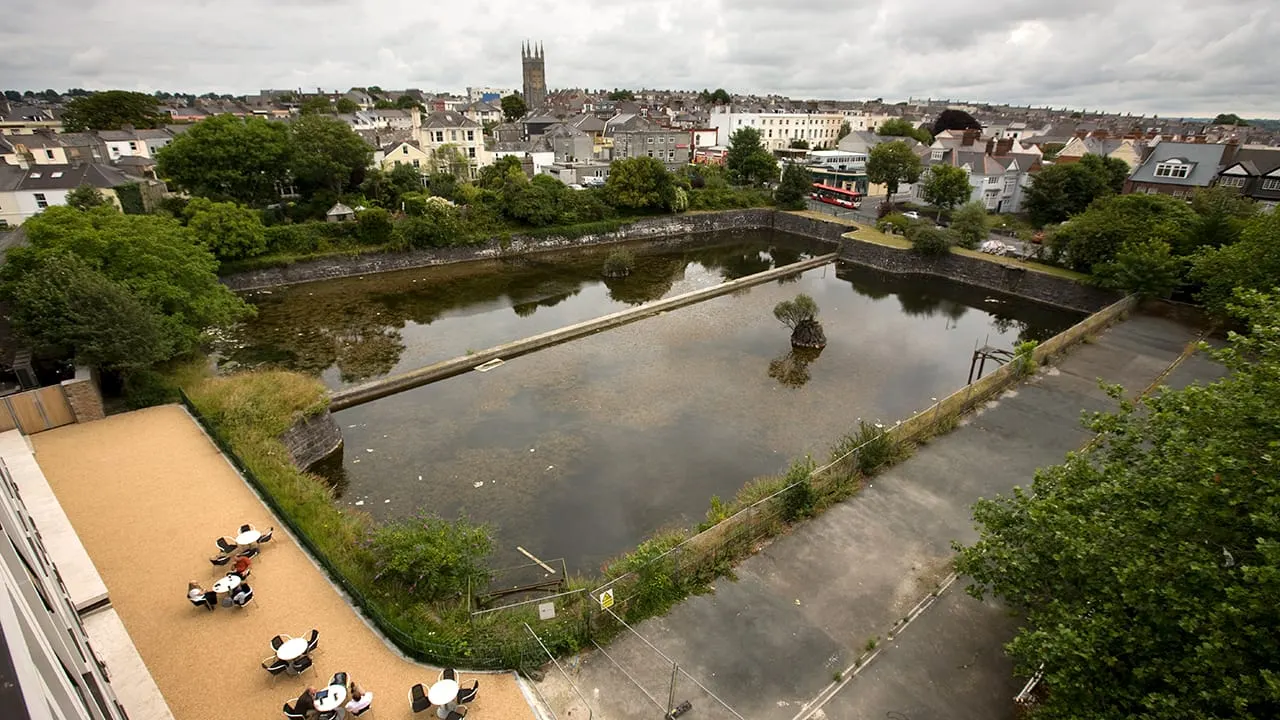 Aerial view of a riverside terrace with visitors relaxing, overlooking a waterway and historic town with church spire beyond.
