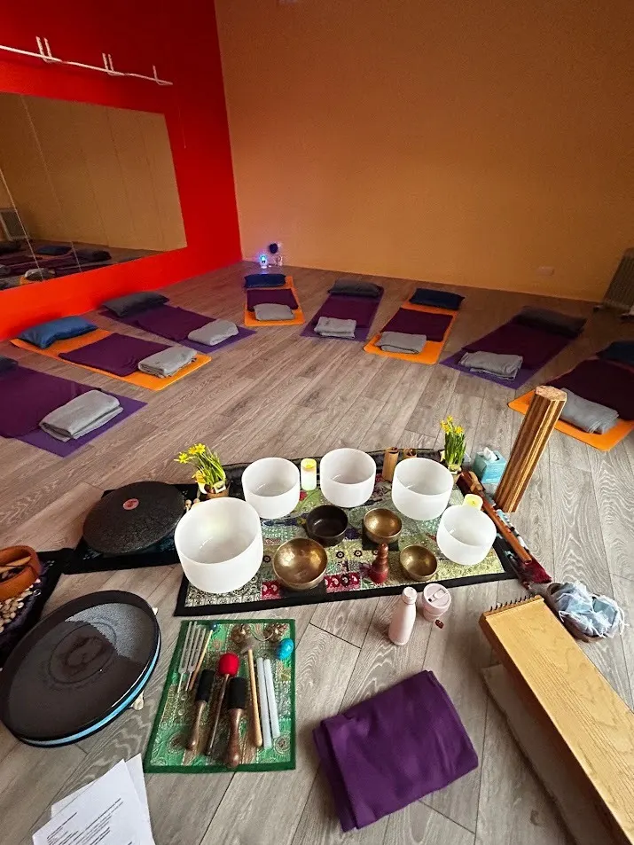 A yoga and wellness studio with purple mats arranged on wooden flooring, singing bowls and meditation instruments displayed in the foreground, and red and gold accent walls.