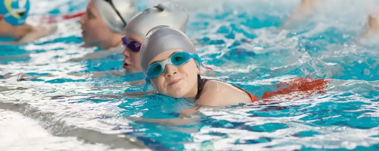 Child swimming in pool wearing goggles and swimming cap, with other swimmers in the background.
