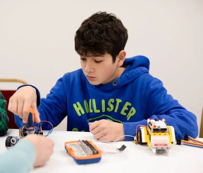 Child in blue hoodie building a robot with electronic components and yellow wheeled chassis at a table.