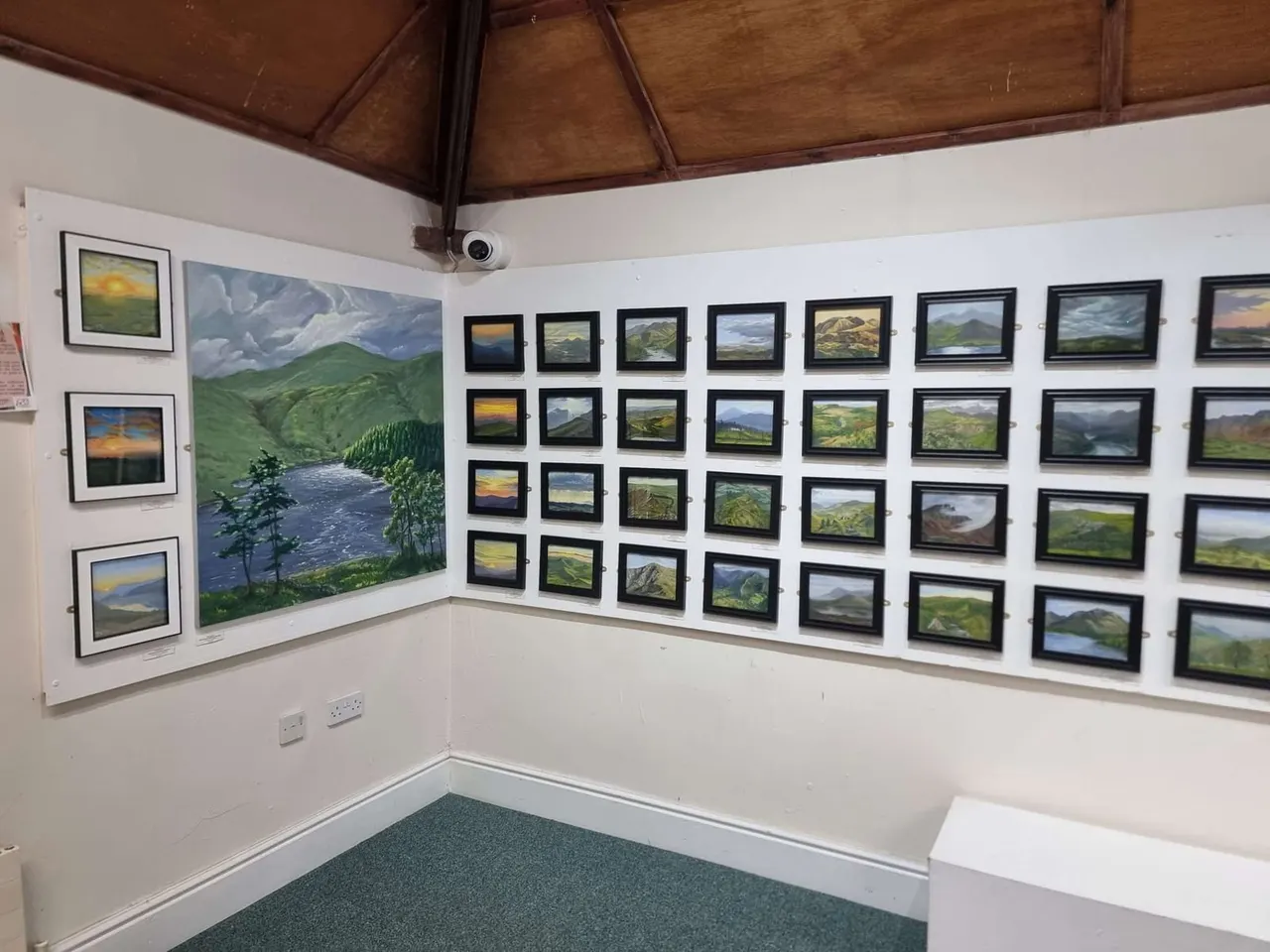 Gallery room with white walls displaying framed landscape paintings and photographs of mountains, valleys and rivers.
