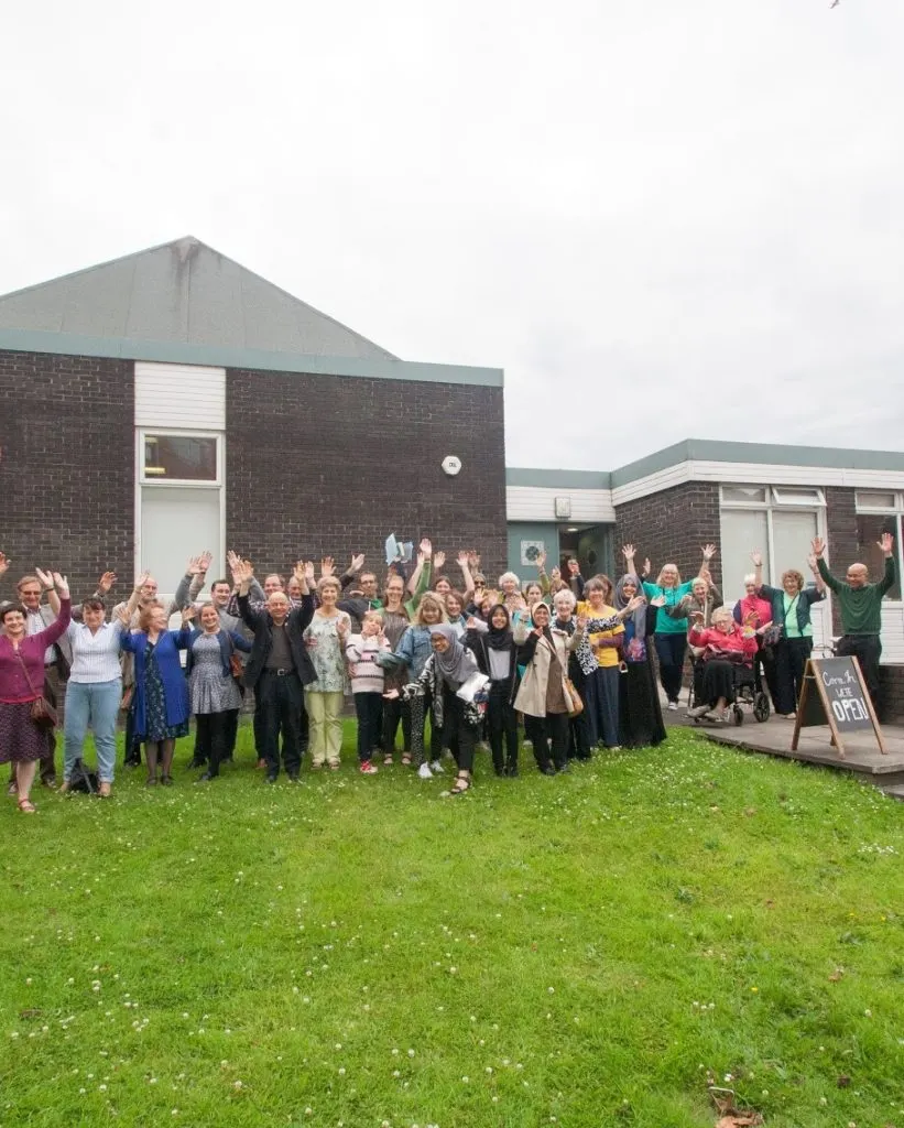 Large group of people of various ages waving at the camera outside a brick community building.