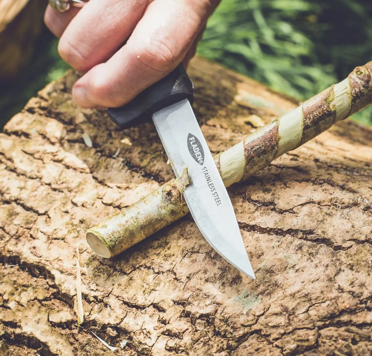 Person using a bushcraft knife to cut bark from a branch outdoors.