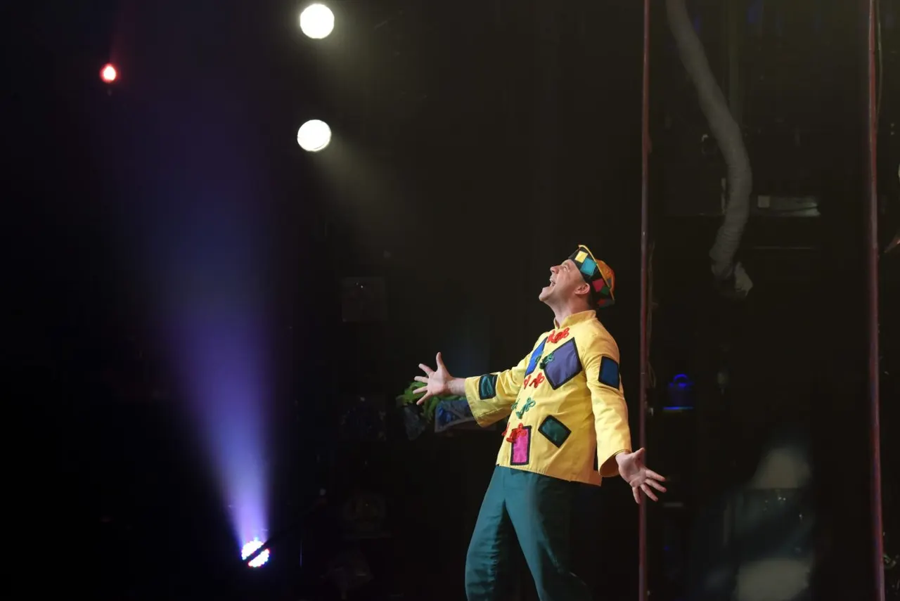 A clown in a bright yellow jacket and colourful hat performs on stage, juggling whilst looking upward under theatrical spotlights.