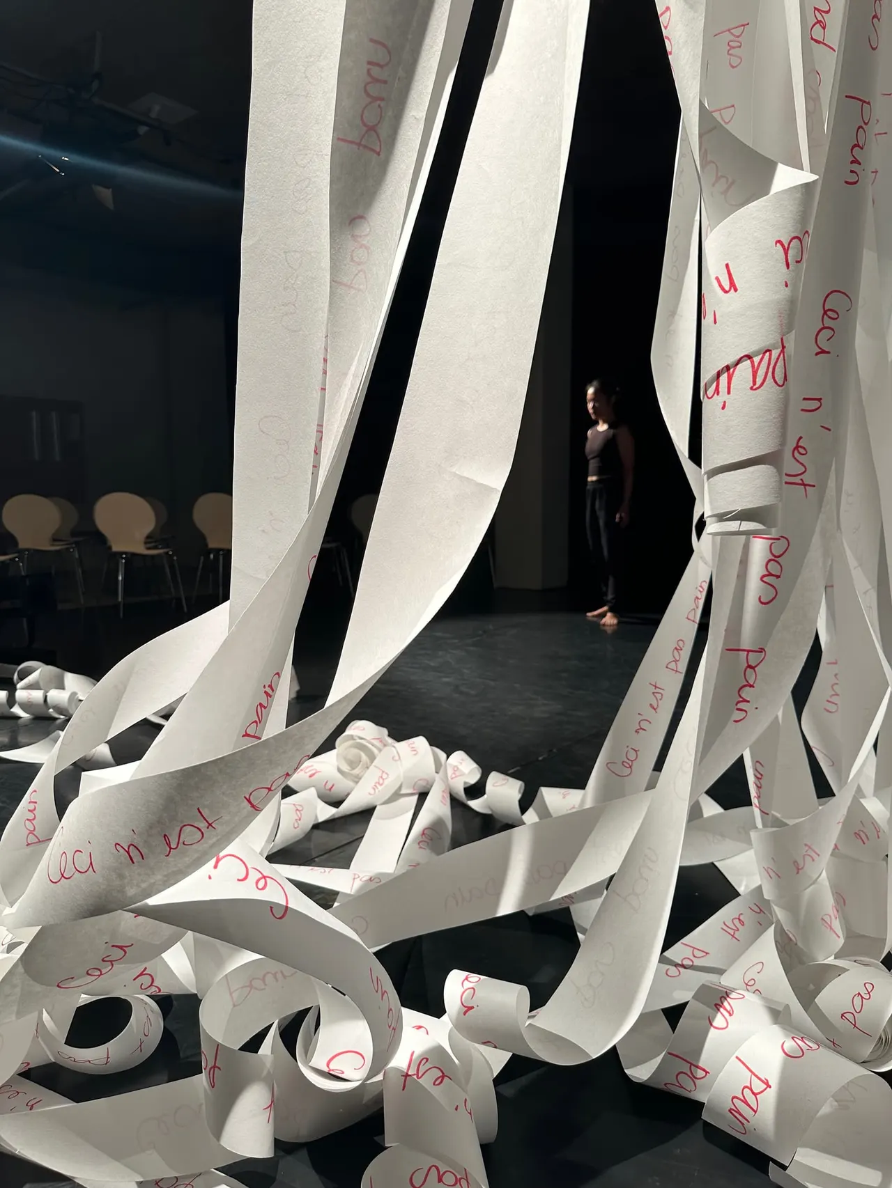 A person stands within an interactive art installation made of white paper ribbons with red handwritten text, inside a contemporary gallery space.