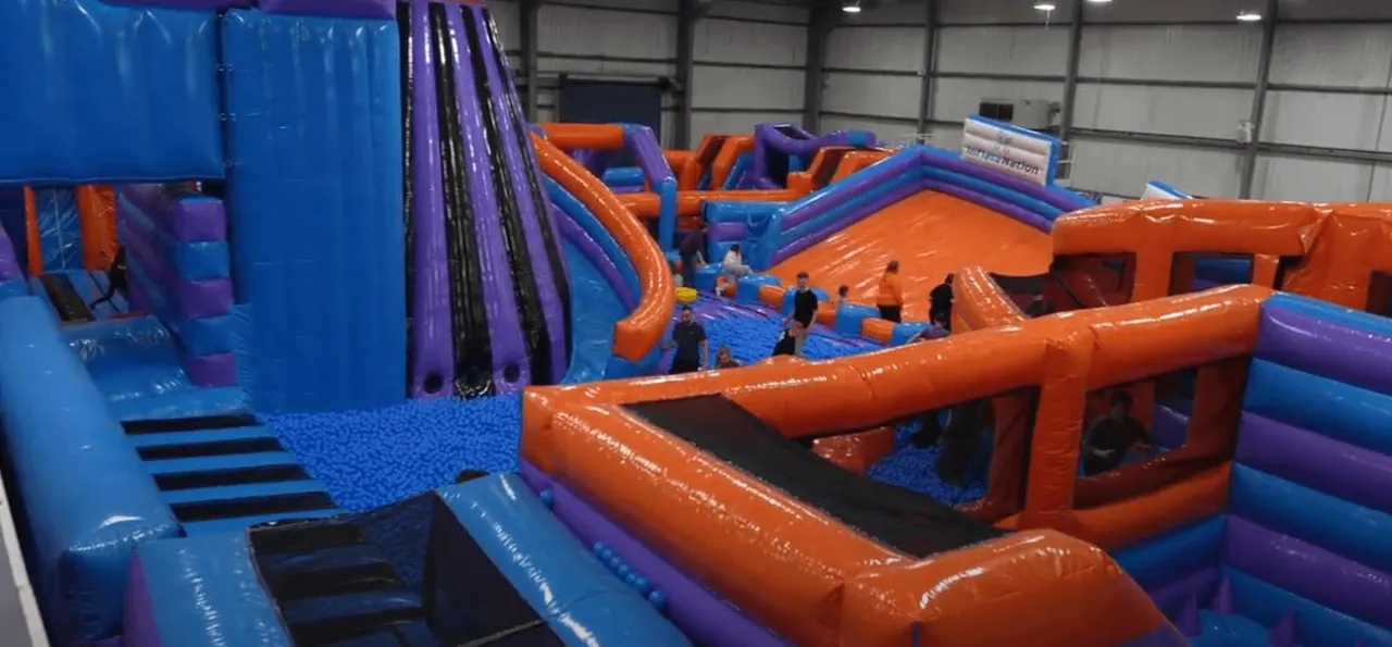Children playing on large inflatable obstacle course structures in bright orange and blue colours inside an indoor venue.