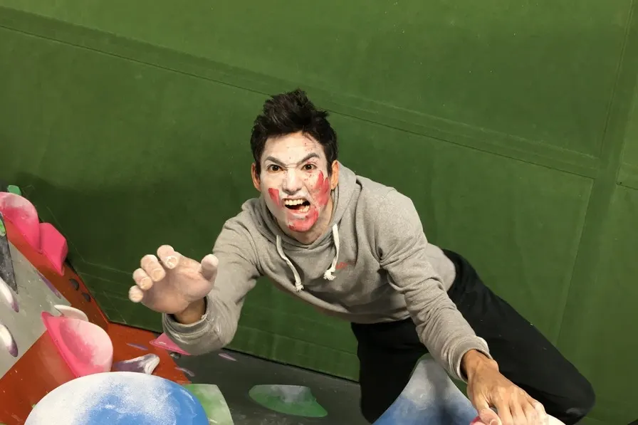 Person with face paint wearing a grey hoodie climbing indoors against a bright green wall with coloured handholds.