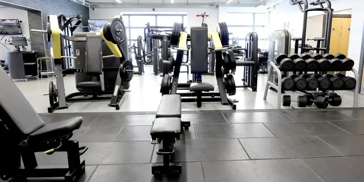 Modern gymnasium interior with weight machines, dumbbells, benches, and barbells arranged throughout the spacious facility.