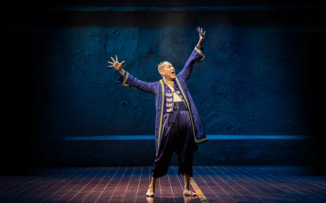 A male performer in an ornate purple and gold costume stands on stage with arms raised triumphantly against a blue textured backdrop.