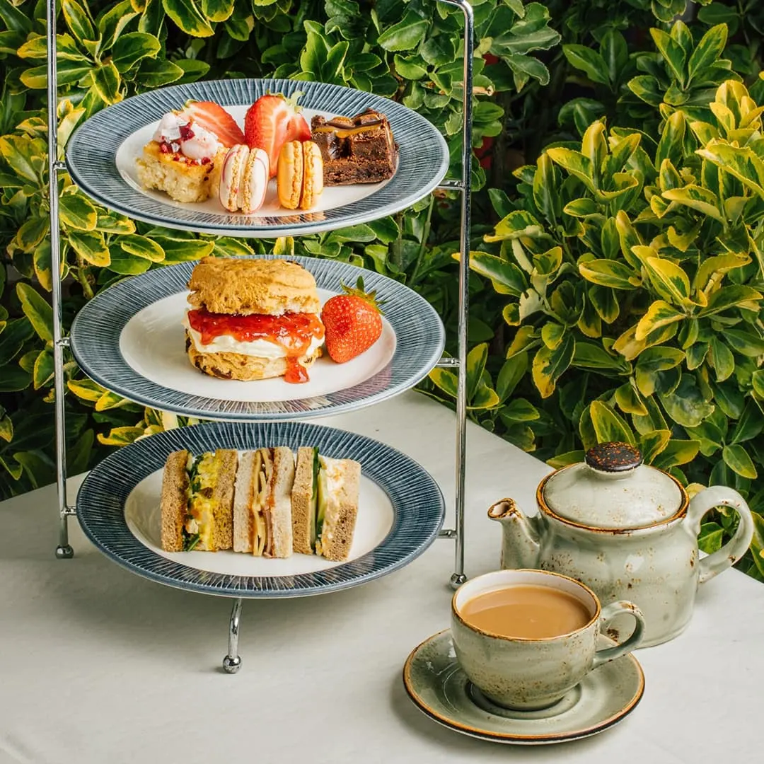 Three-tiered stand displaying afternoon tea with sandwiches, scones, cakes and strawberries, beside a teapot and cup against a green foliage backdrop.