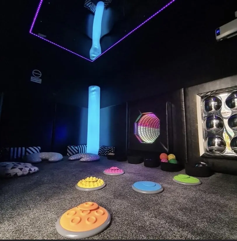 A sensory play room with neon lighting, illuminated floor panels in various colours, textured play mats, and interactive light-up displays on the walls and ceiling.