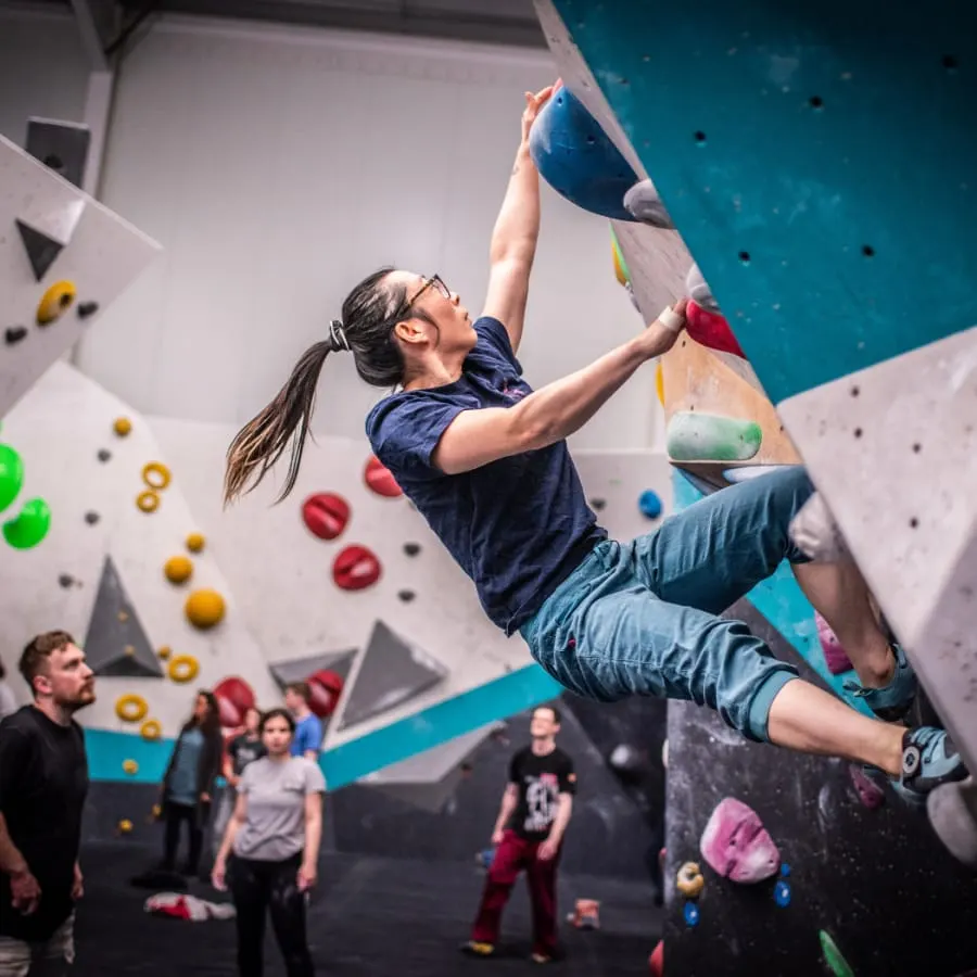 Woman indoor rock climbing on a turquoise and white wall whilst spectators watch below.