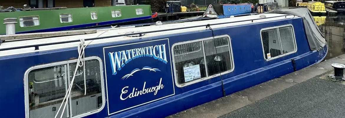 Blue and white narrowboat moored at canal basin with other vessels visible in background.