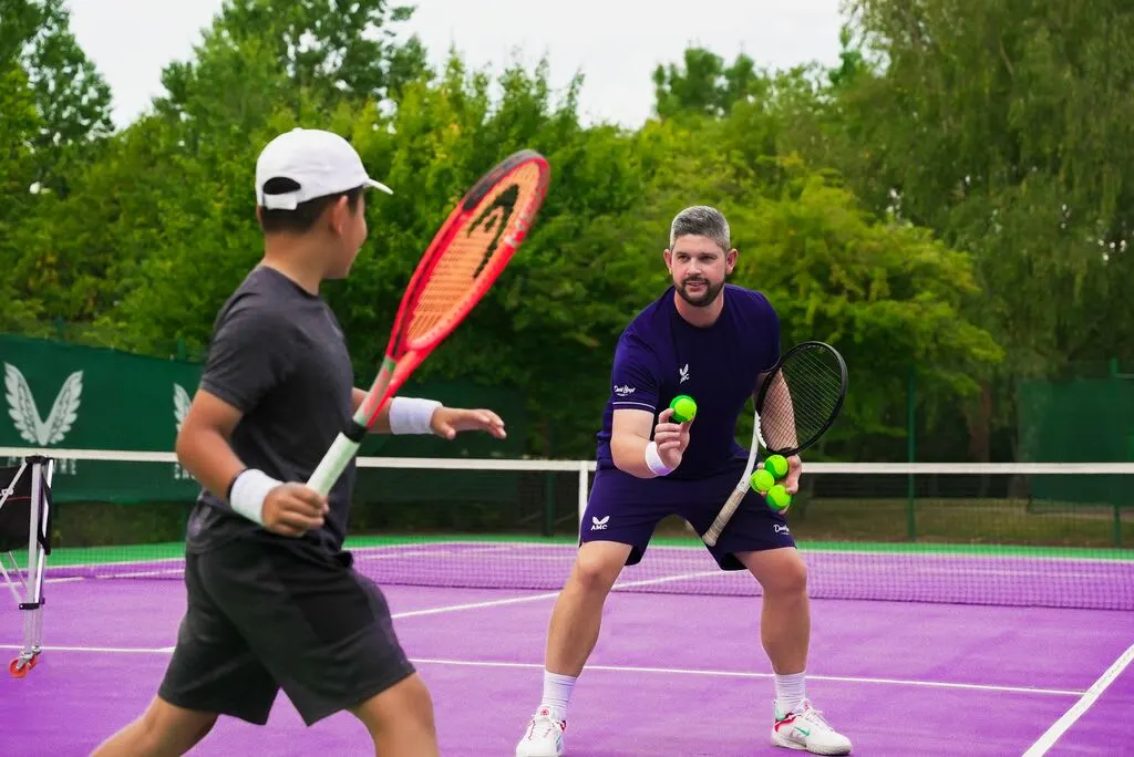 Two male tennis players during a doubles match on a purple court with trees in the background.