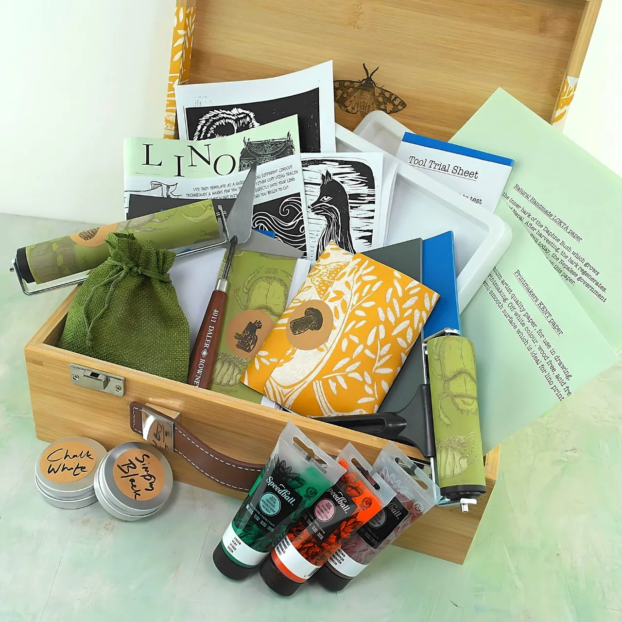 Wooden box of printmaking and art supplies including tools, instruction sheets, paints, and printed artwork samples.