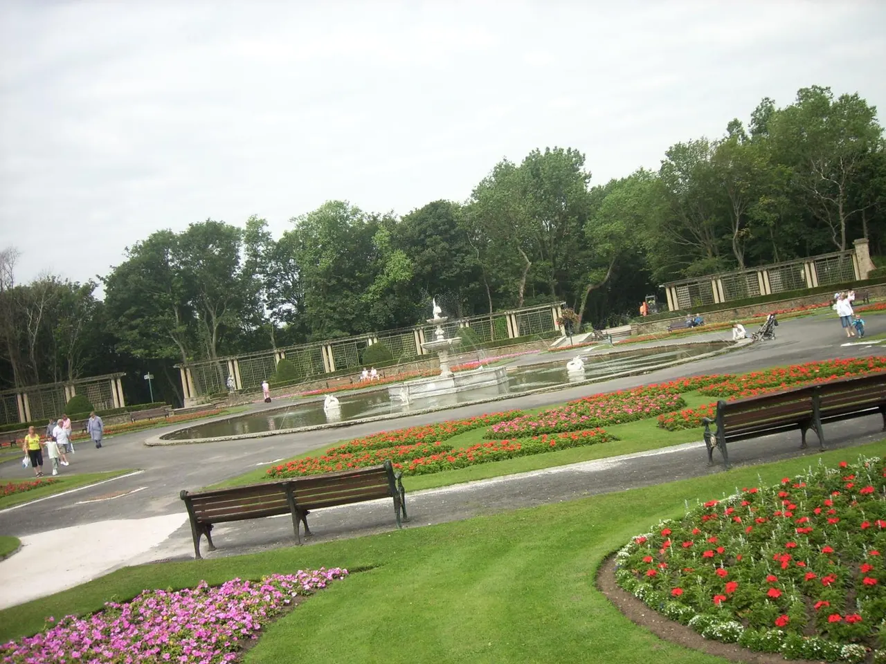 Formal gardens with red and pink flower beds, benches, a central pond with fountain, and visitors walking amongst tree-lined pathways.
