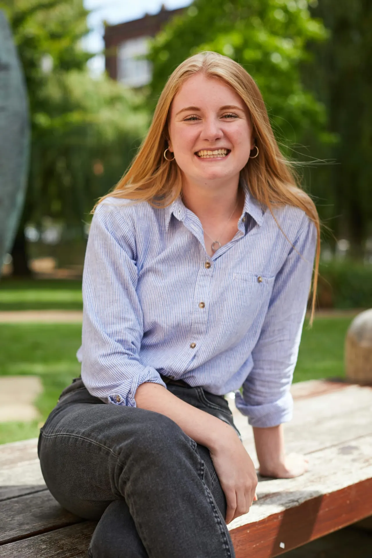 Woman with long reddish hair smiling at camera whilst kneeling on wooden bench in sunny garden setting.