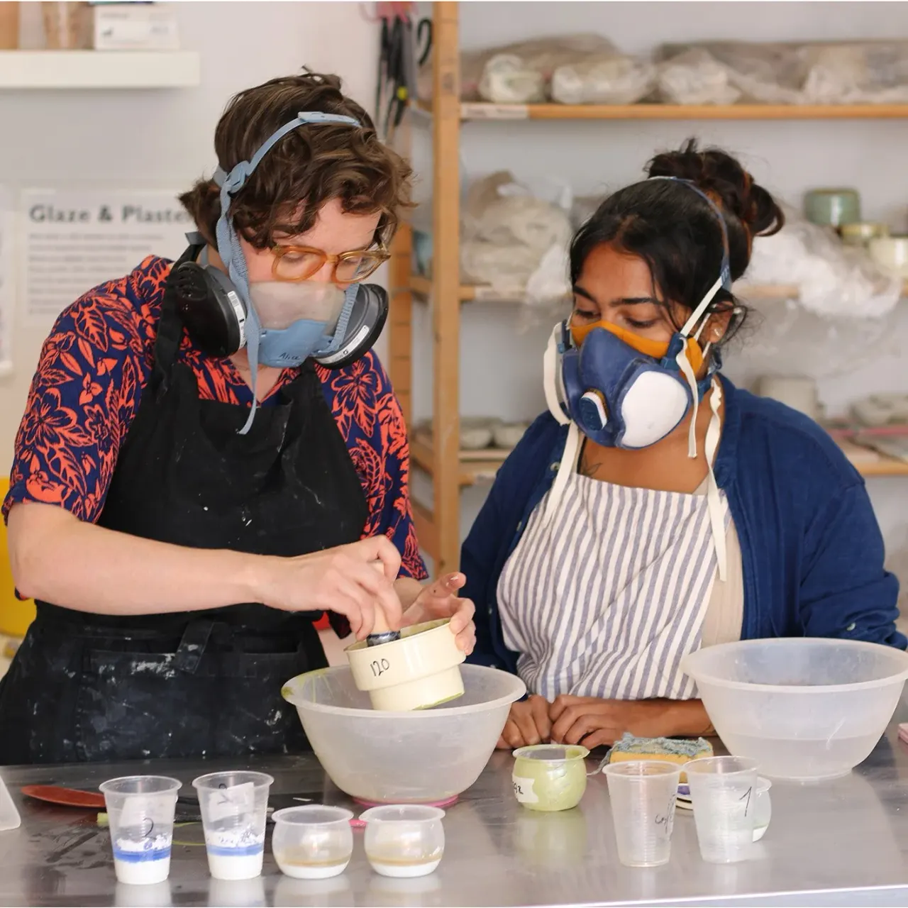 Two people wearing safety masks and headphones work together at a pottery table, mixing glazes in bowls and small containers.