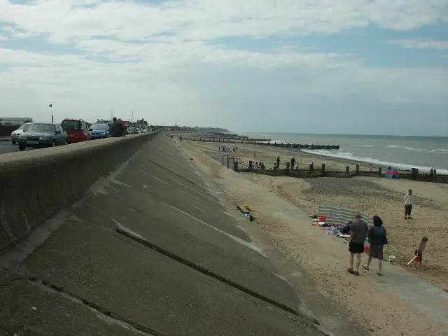 Beach visitors relaxing and swimming on sand beneath a concrete sea wall with parked vehicles above.