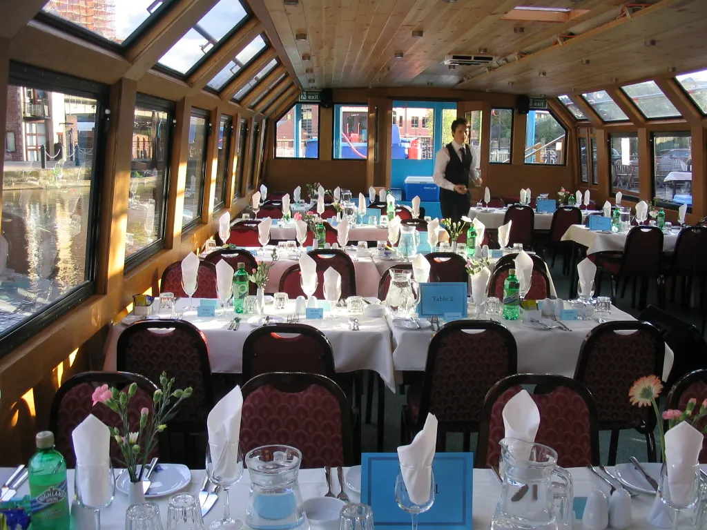 Formal dining room on a river boat with burgundy chairs, white tablecloths, place settings, and floral centrepieces, with staff member standing and windows overlooking waterside.