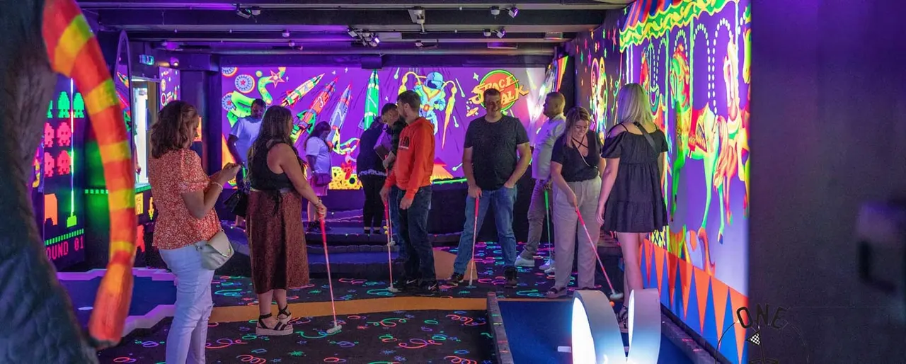Group of people playing mini golf in a neon-lit indoor venue with colourful walls and fluorescent decorations.