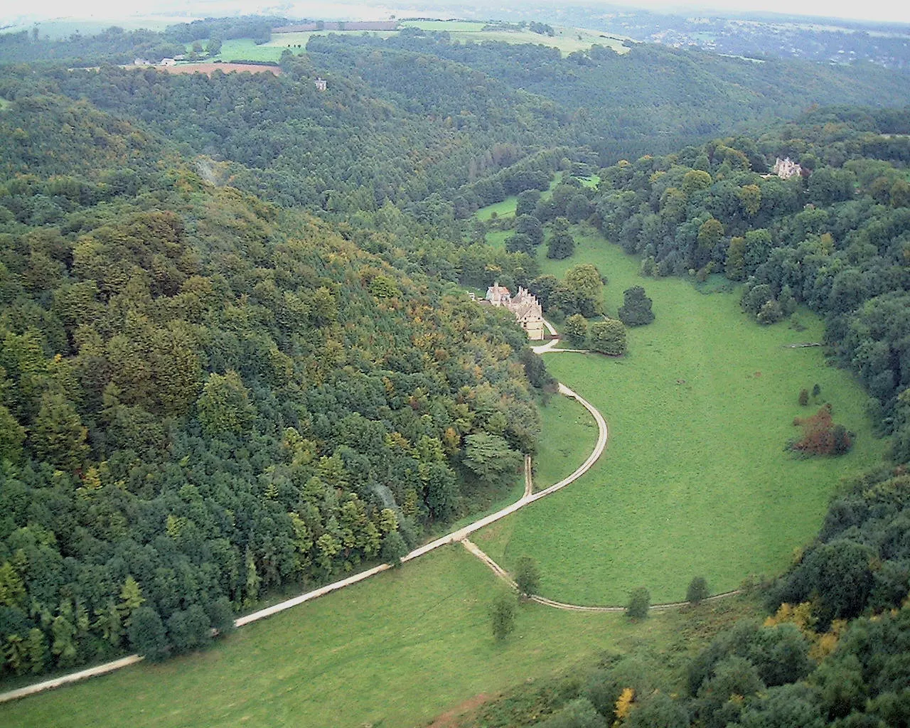 Aerial view of a castle nestled in a wooded valley with winding driveway through green fields and surrounding forest.