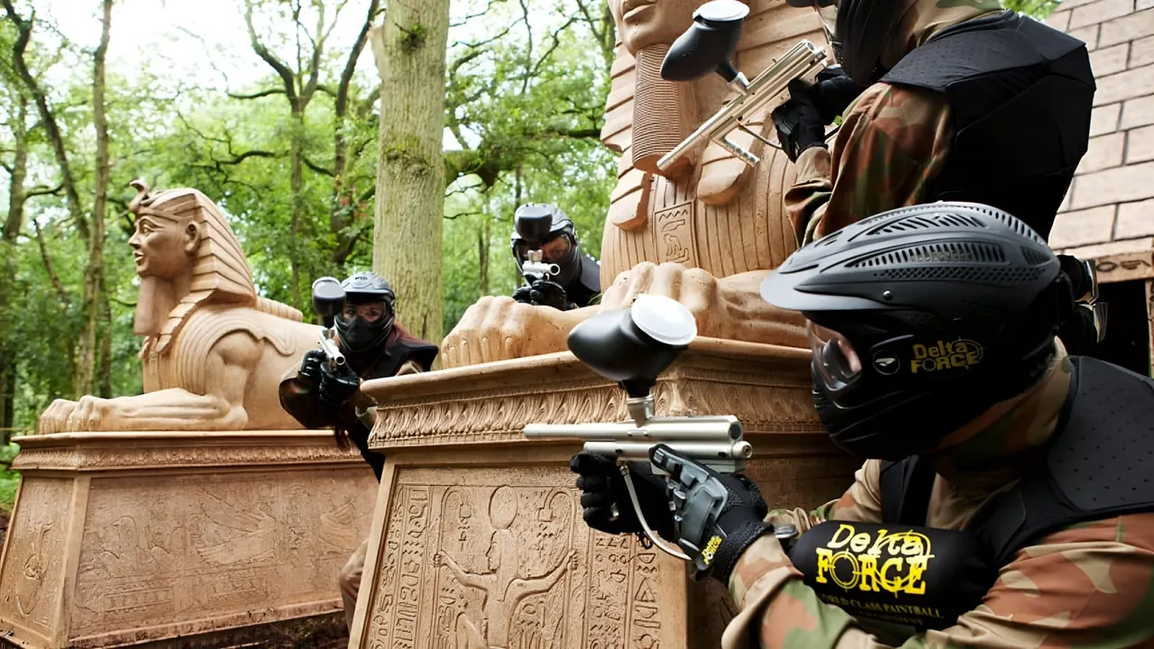 Players in paintball gear with markers aim at Egyptian-themed wooden structures during an outdoor woodland paintball game.