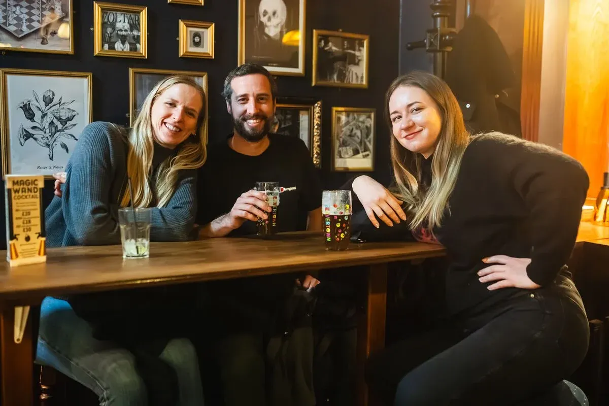 Three smiling people enjoying drinks at a bar counter, with framed artwork and gothic-style décor on the dark walls behind them.