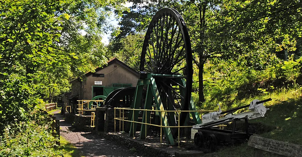 Historic mining equipment including a large water wheel and dark wooden buildings surrounded by dense green woodland.