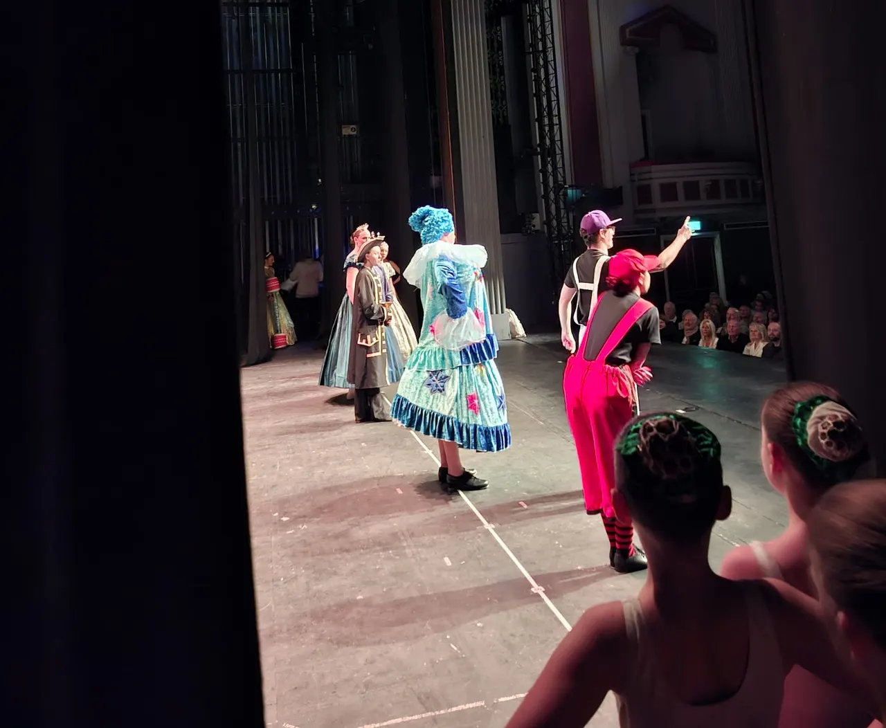 Performers in colourful costumes on an indoor theatre stage, with audience members visible in the foreground and seating area.