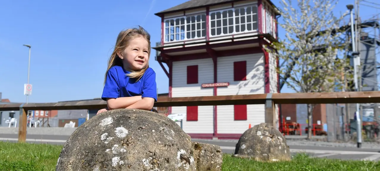 Young girl in blue shirt posing on a large boulder with a heritage colliery building behind her on a sunny day.