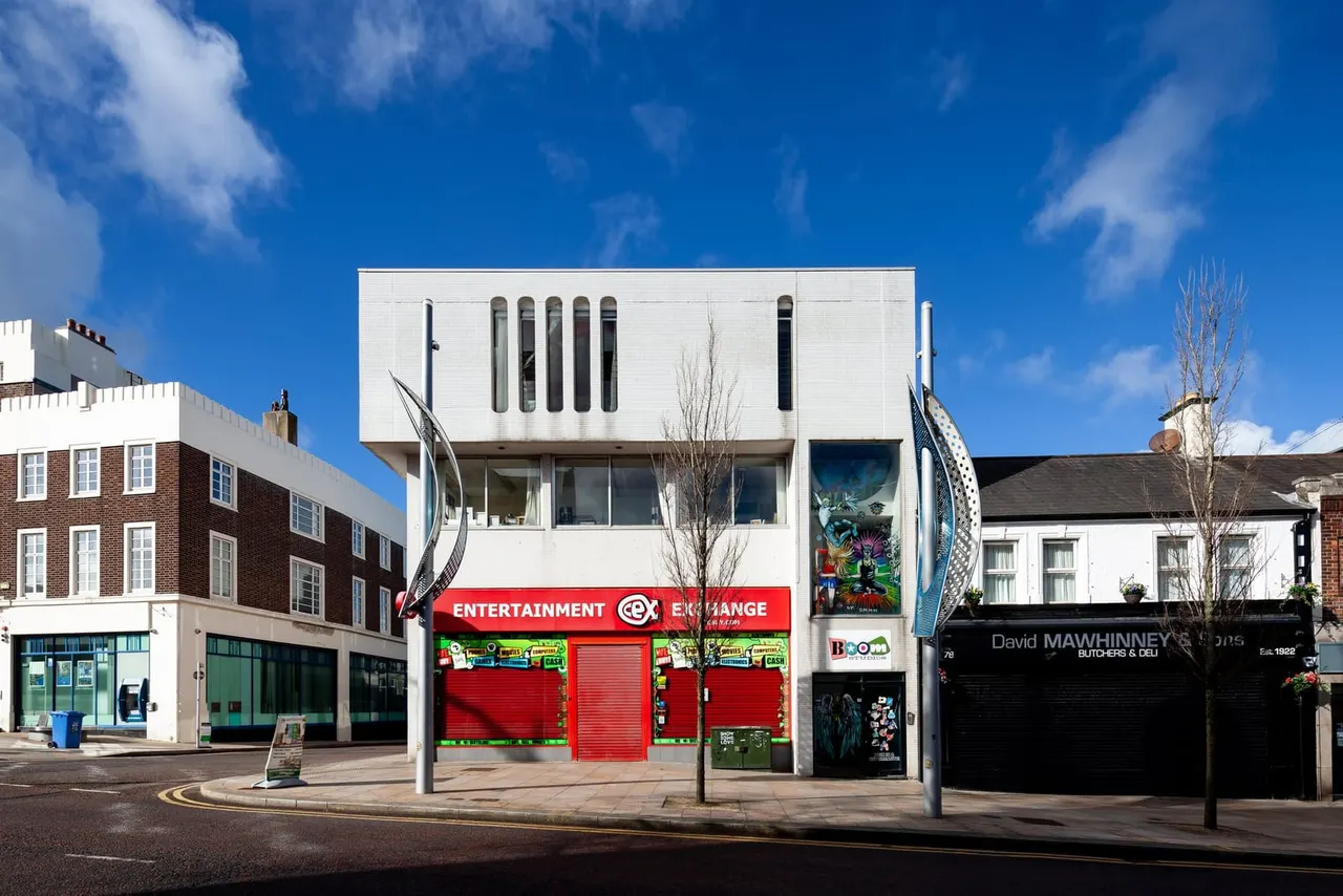 A modernist white building with red-shuttered entertainment venue storefront on a sunny high street surrounded by older brick buildings.